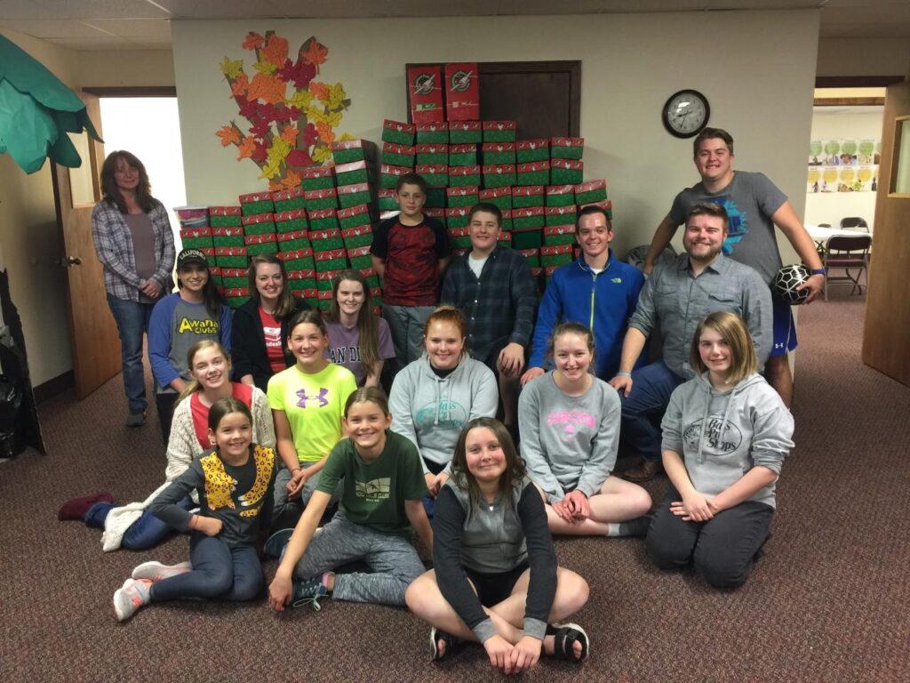 group of kids pose for picture in front of large stacks of Operation Christmas Child shoeboxes
